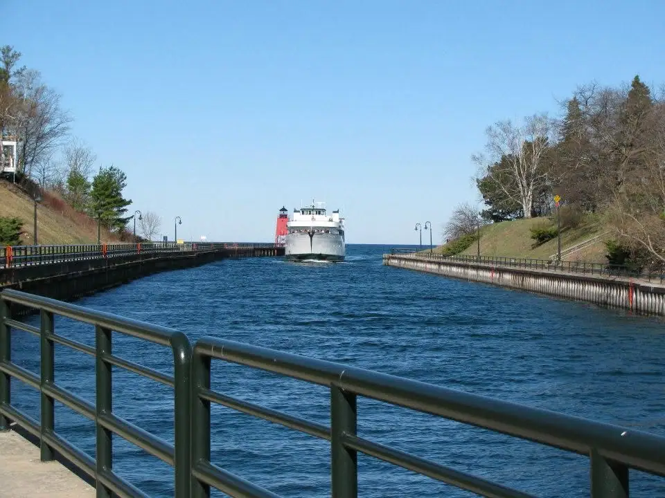 A ferry approaches through a narrow waterway lined with railings, trees, and a red lighthouse tower.