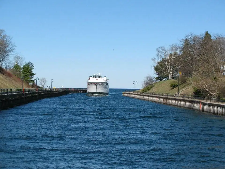 A white ferry approaches through a narrow, walled water channel lined with railings and lampposts.