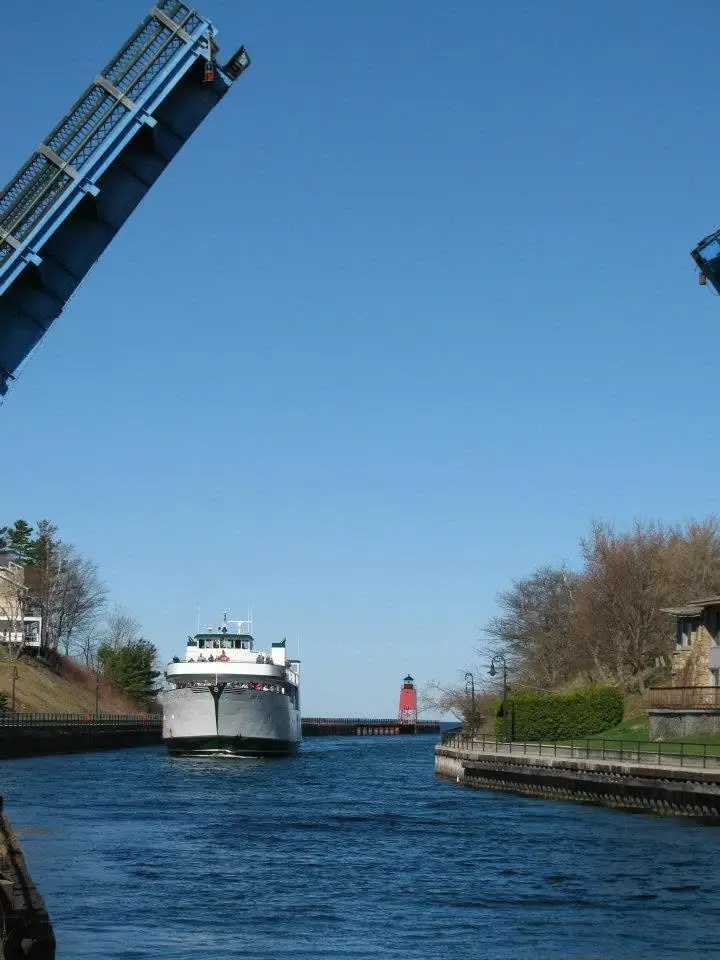A ferry passes beneath a raised drawbridge toward a red lighthouse at the end of a pier.