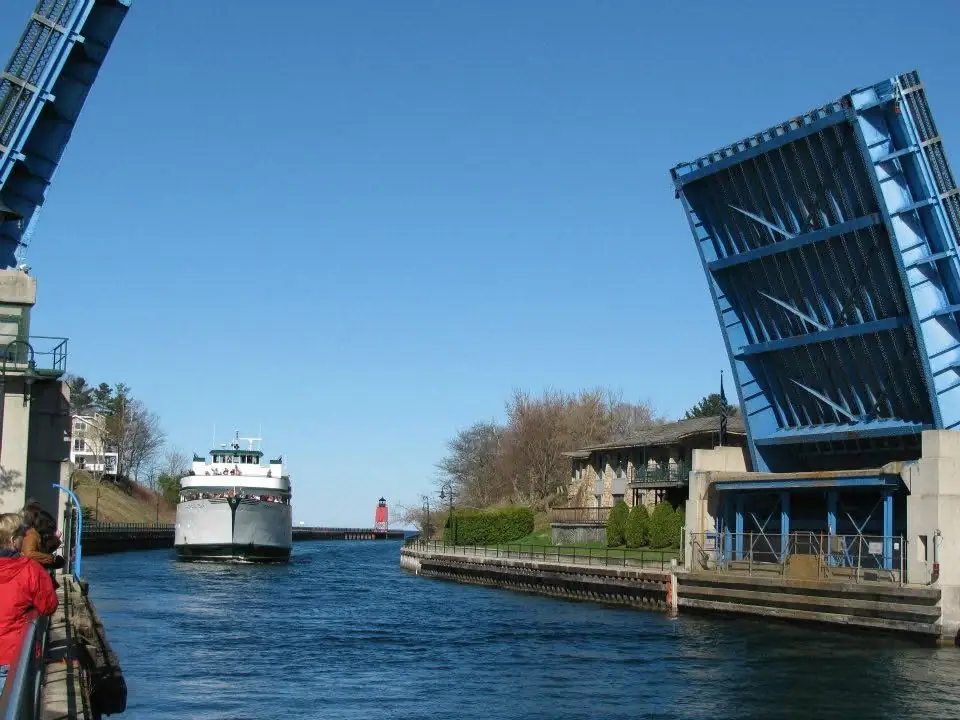 A white ferry passes beneath a raised blue drawbridge toward a red lighthouse.