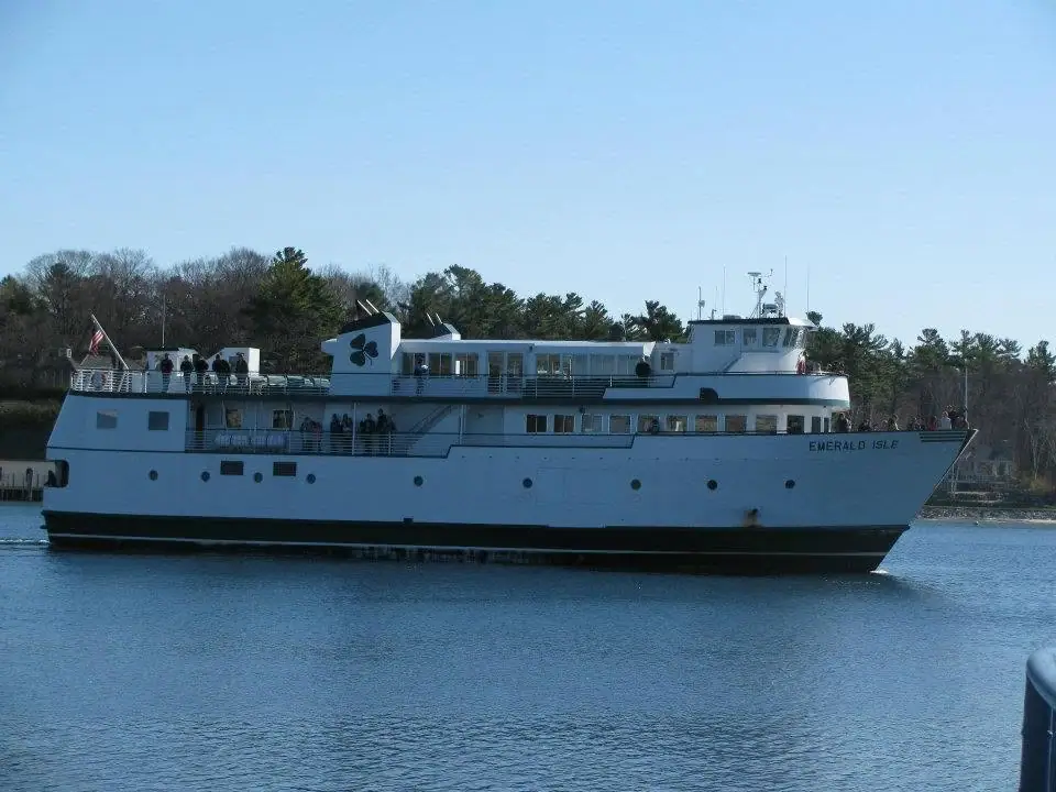 A white passenger ferry with people on deck travels past a wooded shoreline.