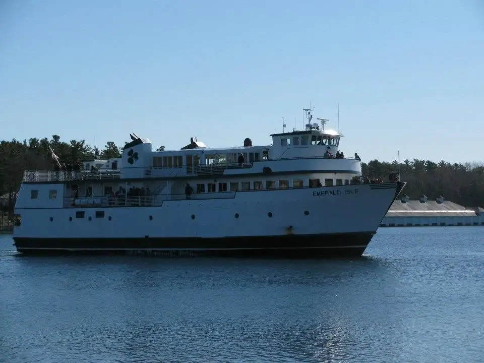 A large white passenger ferry with multiple decks moves across calm water near a wooded shoreline.