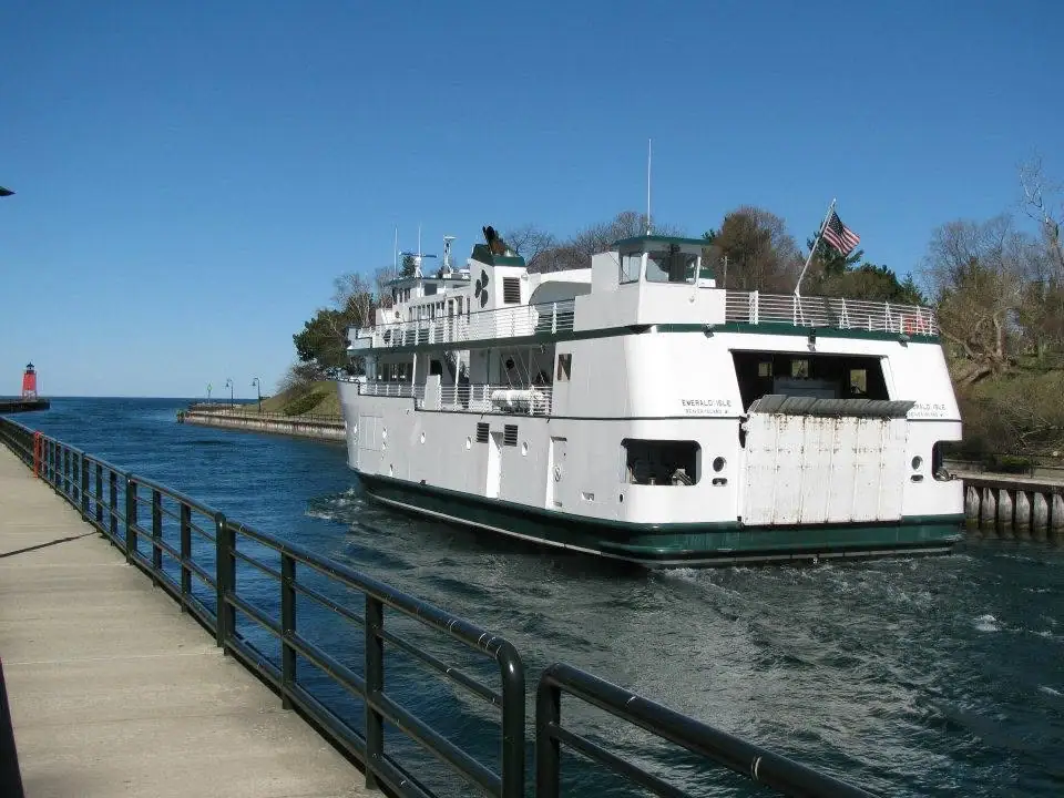 A white ferry passes a pier beside a wooded shoreline, with a red lighthouse visible in the distance.