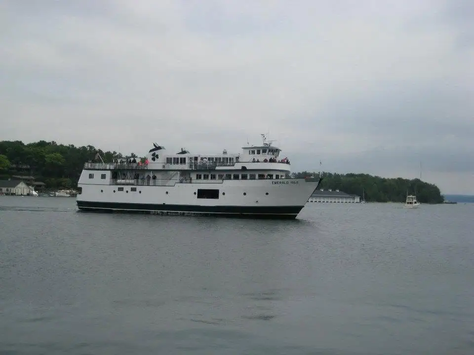 A white multi-deck ferry carrying passengers passes a wooded shoreline under an overcast sky.