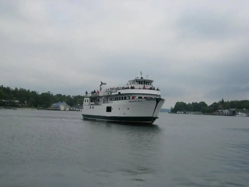 A white passenger ferry carrying people moves through calm water past a tree-lined shore.