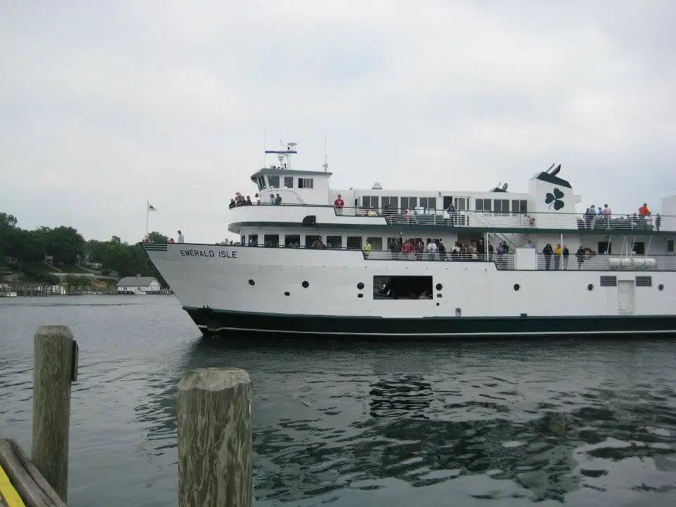 A white passenger ferry carrying people passes wooden dock pilings near a wooded shoreline.