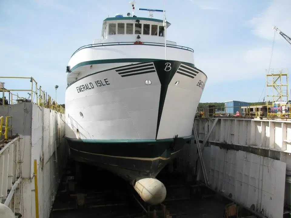 Front view of a white ferry resting in a concrete dry dock, with cranes and industrial equipment around it.