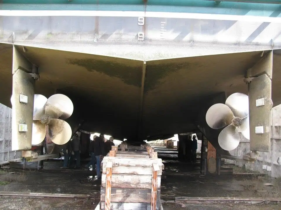 View beneath a large vessel showing twin propellers, support blocks, and people in shadow.