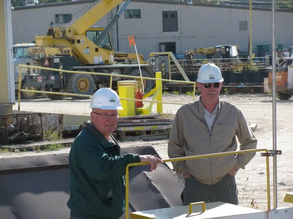 Two hard-hat workers stand beside heavy equipment at an industrial construction site.