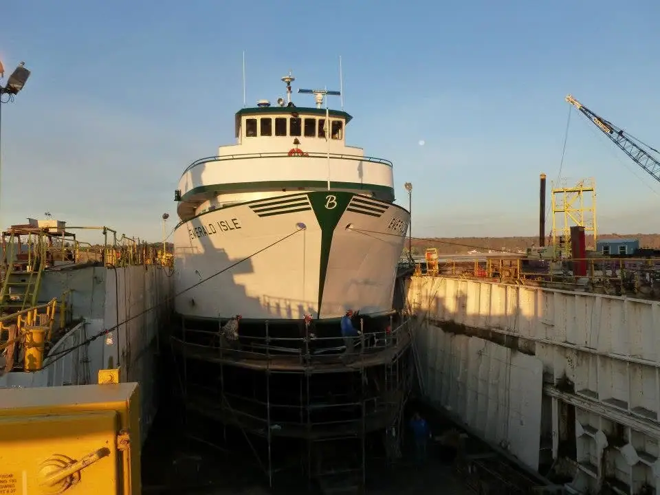 A white ferry sits in a dry dock while workers and a crane surround it in late-day light.