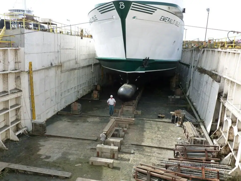 A white-and-green ferry sits in a concrete dry dock above a worker and ship-repair materials.