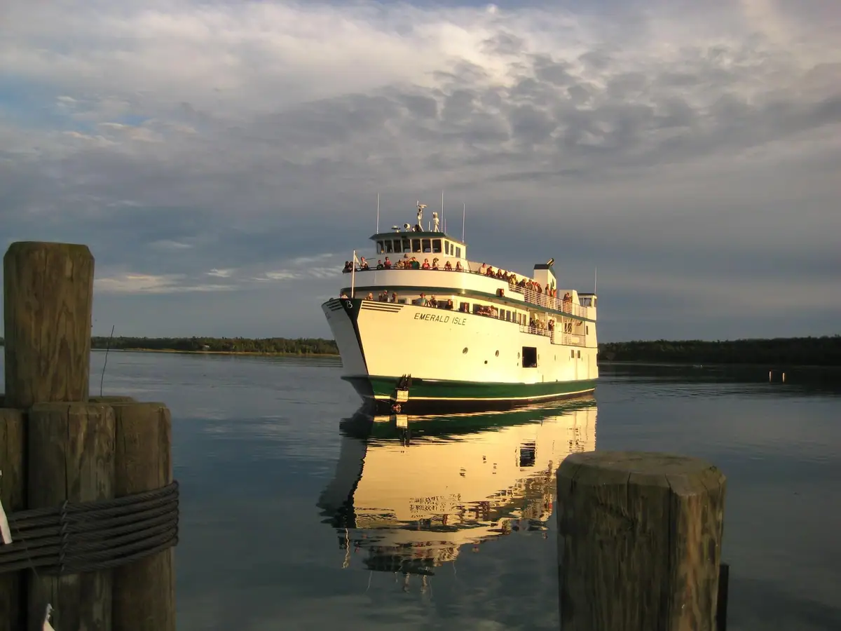 A white passenger ferry approaches wooden pilings on calm water beneath a cloudy sky.