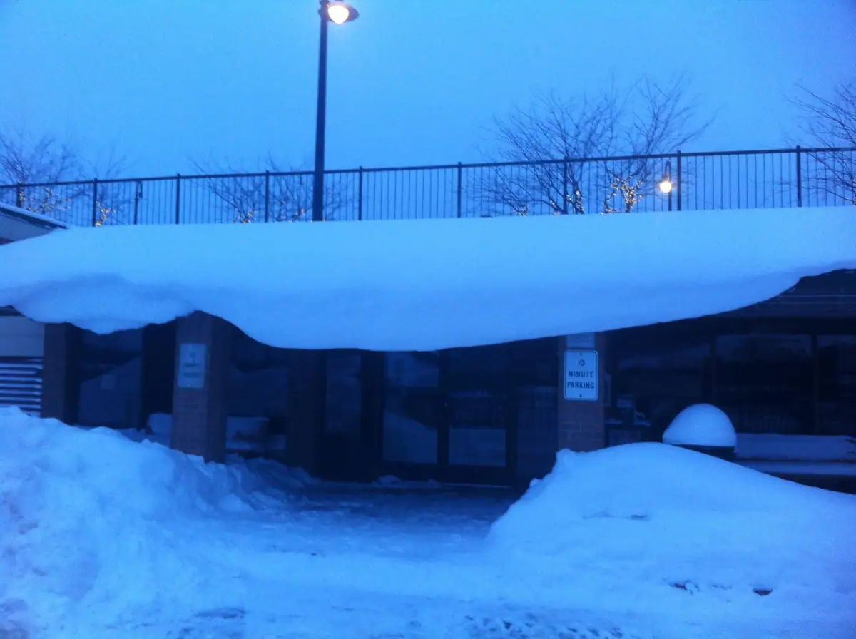 A snow-covered building entrance at dusk, with a parking sign and street lamps.