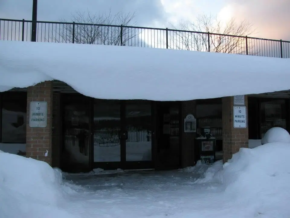 A glass-door building entrance is surrounded by deep snow, with parking signs on brick pillars.