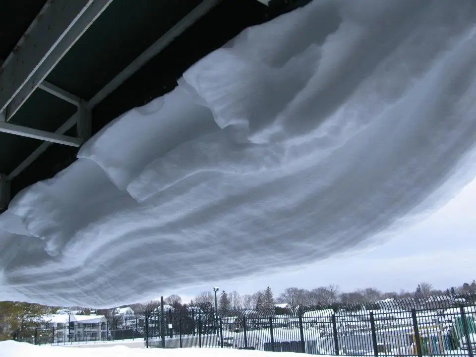 A broad, layered sheet of ice and snow hangs beneath a roof above a fenced, snow-covered grounds.