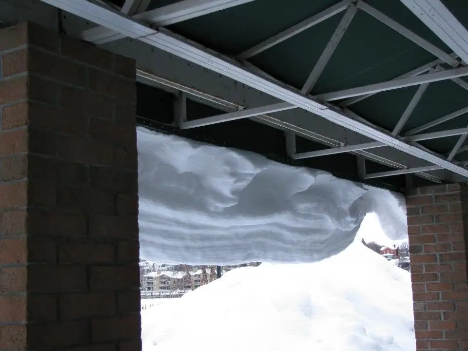 A massive layered snowdrift hangs beneath a roof supported by brick pillars and metal trusses.