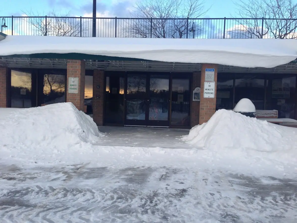 Snowbanks surround a glass-fronted brick entrance with parking signs and a deep layer of snow along the roof.