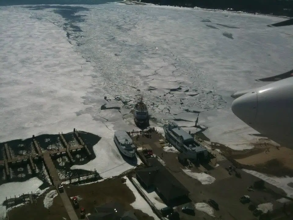 Aerial winter view of ferries, docks, buildings, and broken ice beside an aircraft engine.