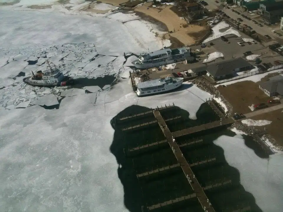 Aerial photograph of an ice-filled harbor with a cutter, two ferries, docks, and snow-covered shorelines.
