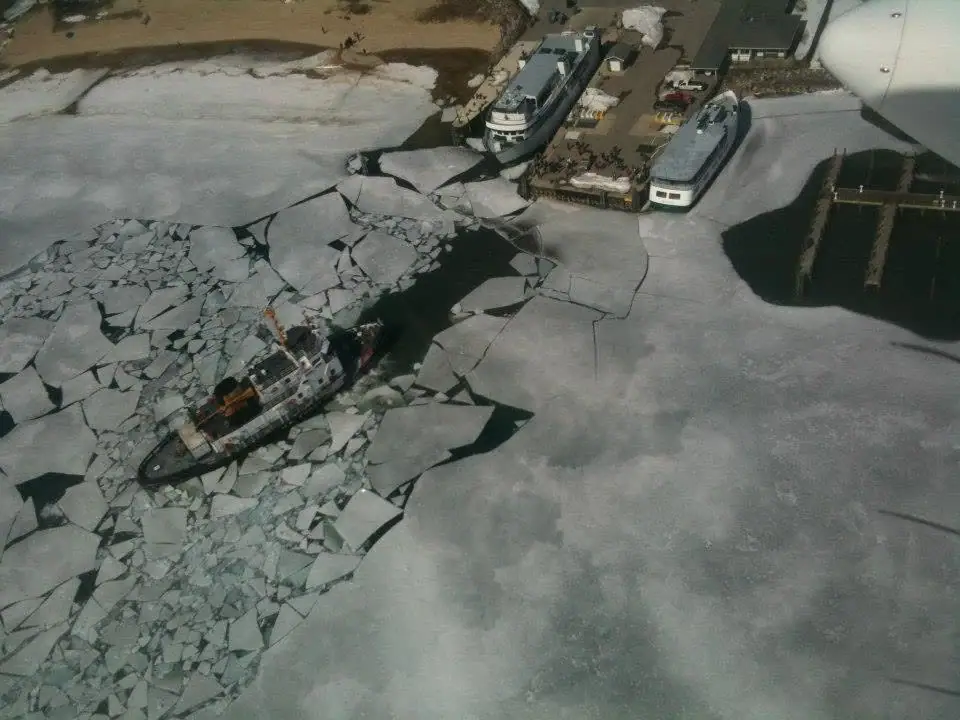 Aerial photograph of a vessel moving through broken ice toward two passenger vessels at a dock.