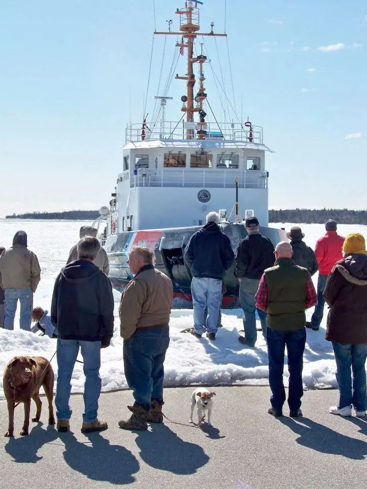 People and two dogs stand on a snowy dock facing a white ferry with a tall mast.