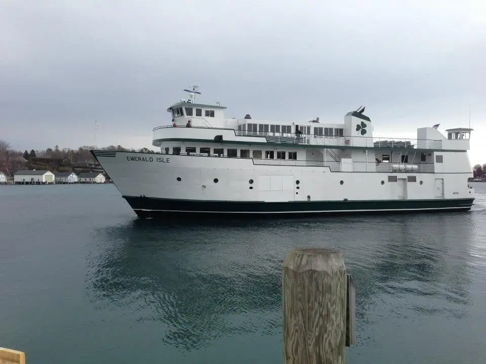 A large white ferry with green trim passes a shoreline, viewed from beside a weathered wooden piling.