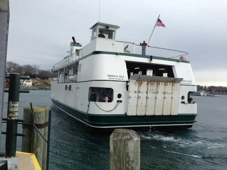 A white-and-green ferry pulls away from a pier, with people aboard and an American flag flying above.