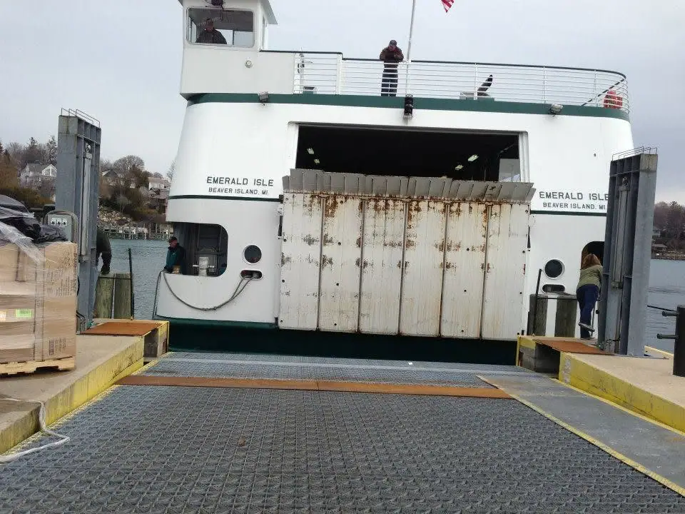 A white ferry faces a dock with its vehicle ramp lowered and people on deck and beside the loading area.