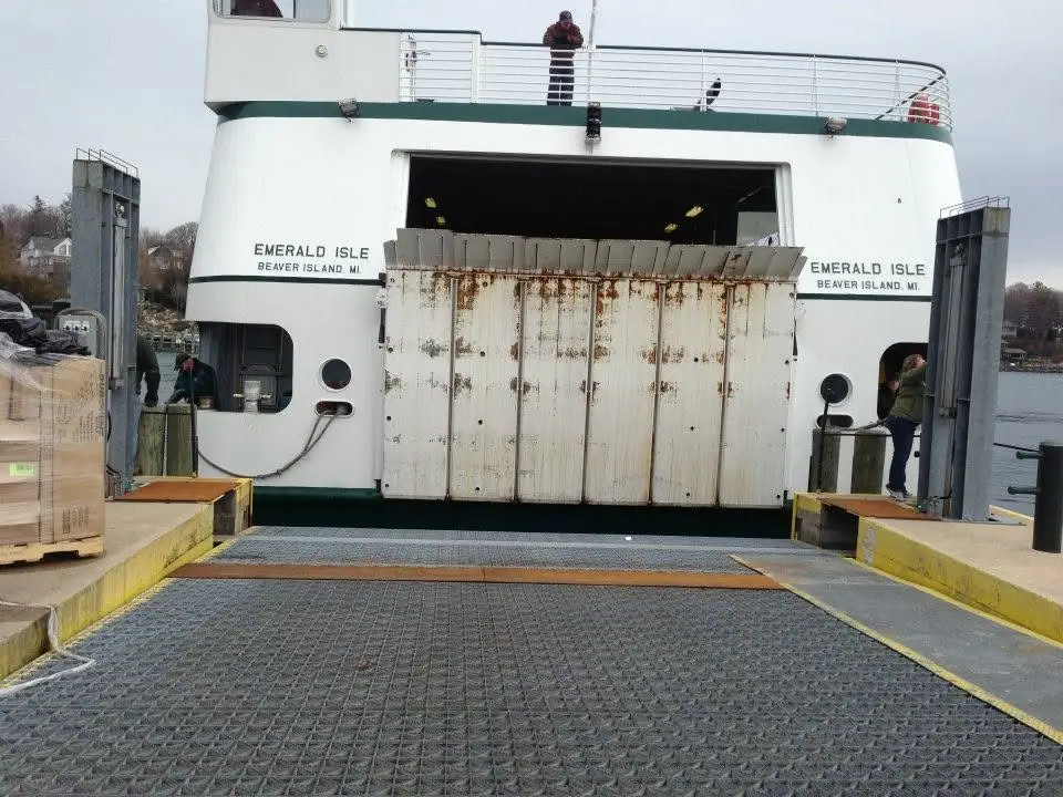 A white ferry is positioned at a loading ramp while cargo is handled at its open stern.