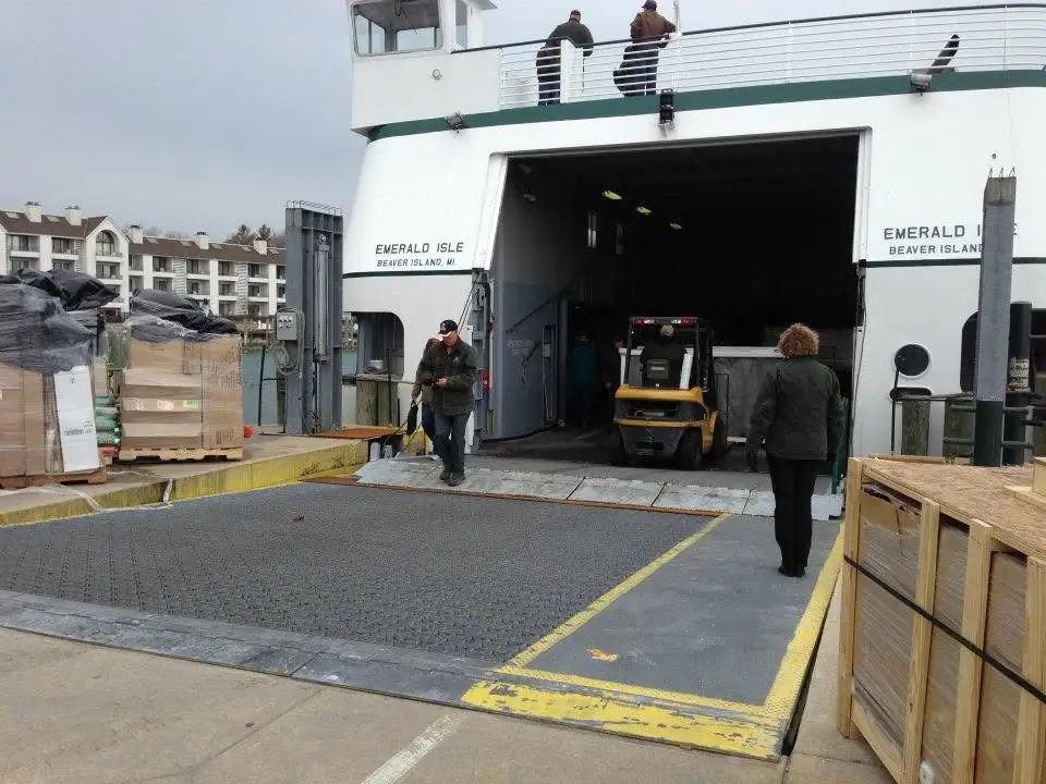 A ferry's open loading bay faces a ramp with a forklift, workers, and palletized cargo.