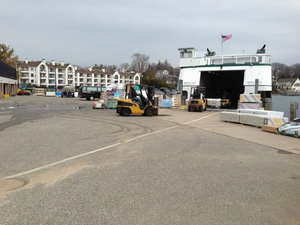 A ferry with an open vehicle deck is loaded at a dock while forklifts move pallets nearby.