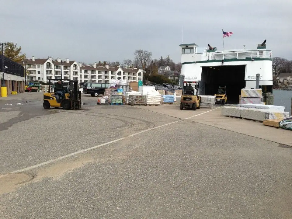 Ferry with an open vehicle deck beside a dock, surrounded by forklifts and stacked pallets.