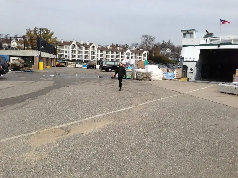 A woman crosses a paved lot beside a ferry, stacked cargo, and apartment buildings.