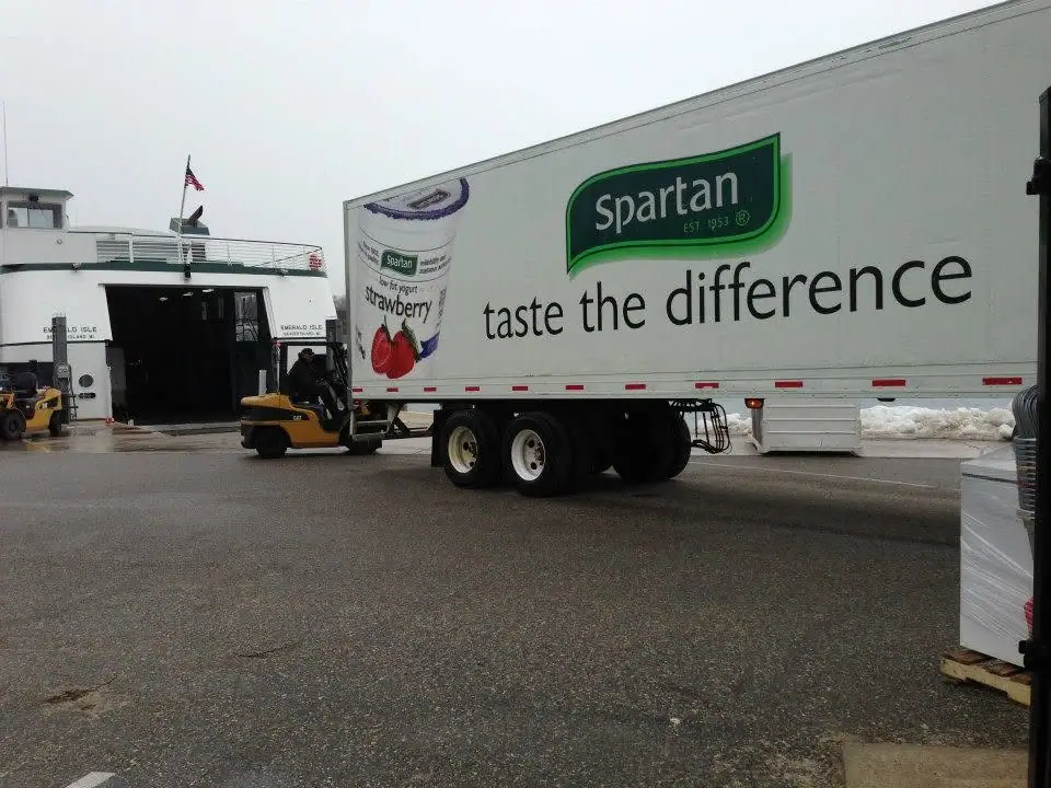 A yogurt advertising trailer stands beside a ferry while a forklift operates in a snowy loading area.