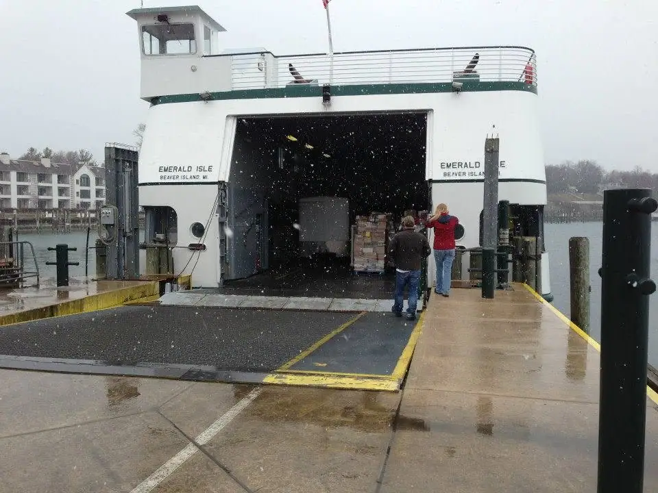 Ferry loading cargo at a wet dock as snow falls, with two people on the ramp.