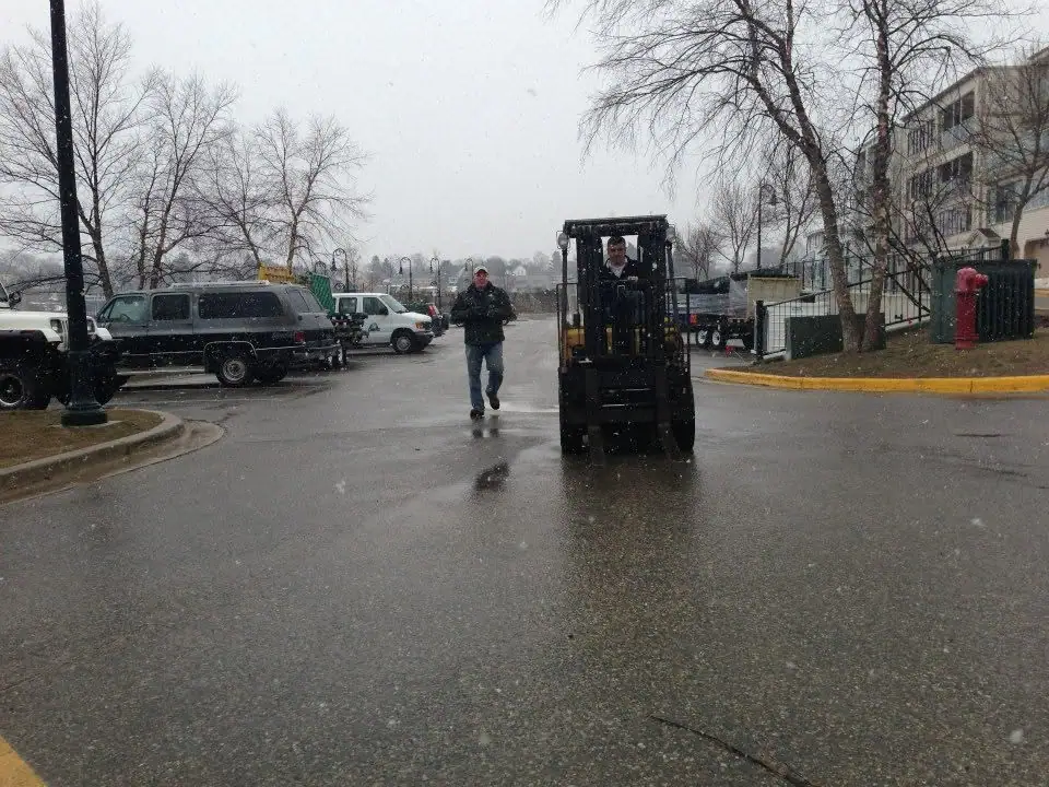 A man walks beside a forklift in a wet, snowy parking lot as light snow falls.