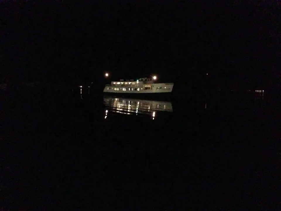 An illuminated passenger ferry moves across dark water at night, with its lights reflected below.