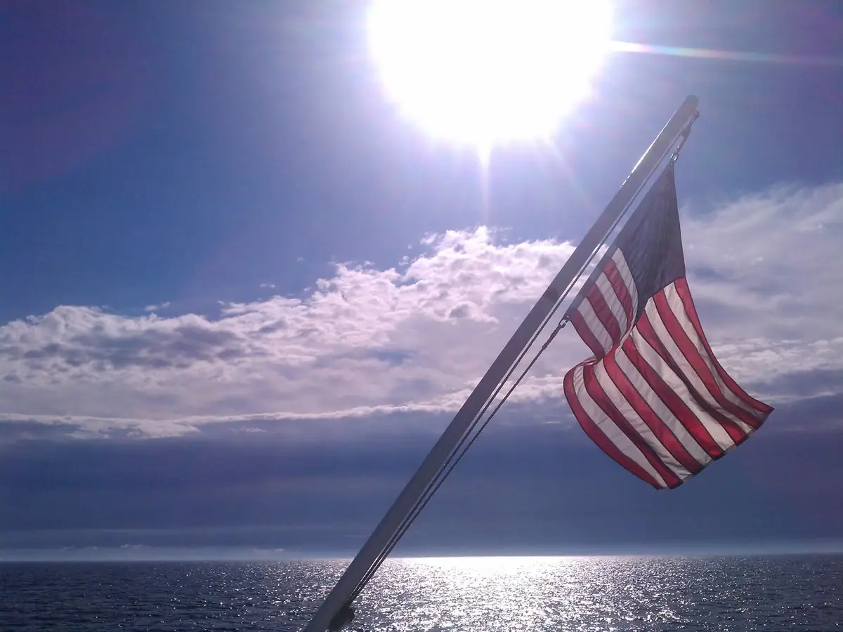 American flag hanging from a slanted mast above sunlit water and a cloudy horizon.