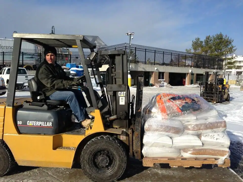 A worker drives a yellow forklift carrying a shrink-wrapped pallet through a snowy outdoor yard.