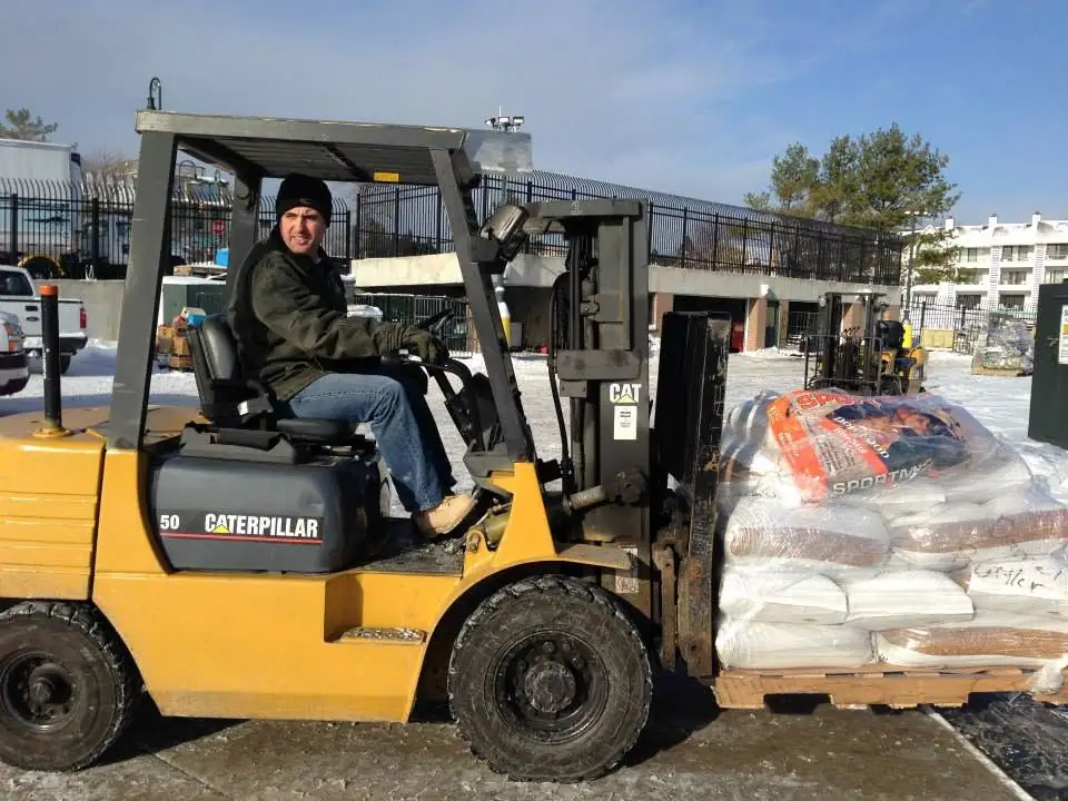A worker drives a yellow Caterpillar forklift carrying a pallet of bagged material through a snowy yard.