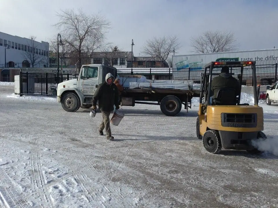 A worker carries buckets across a snowy lot beside a flatbed truck and yellow forklift.