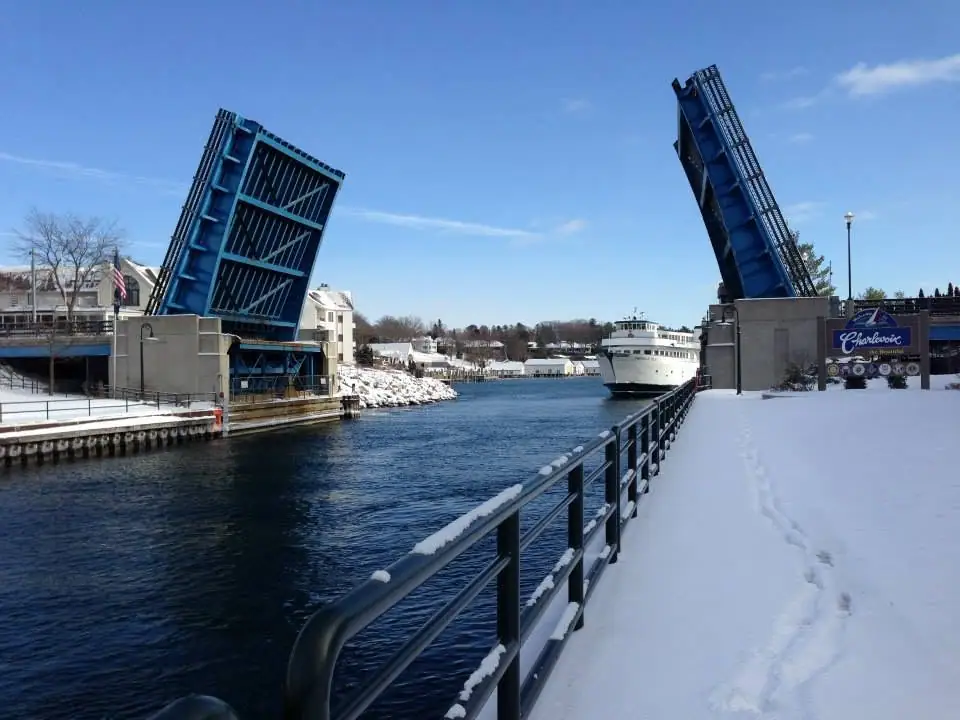A white ferry passes beneath a raised blue drawbridge in a snow-covered waterfront scene.