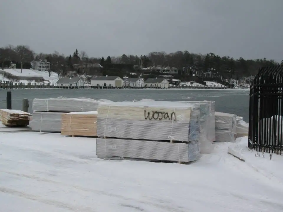 Snow-covered stacks of wrapped lumber sit beside a waterfront, with houses and trees across the water.