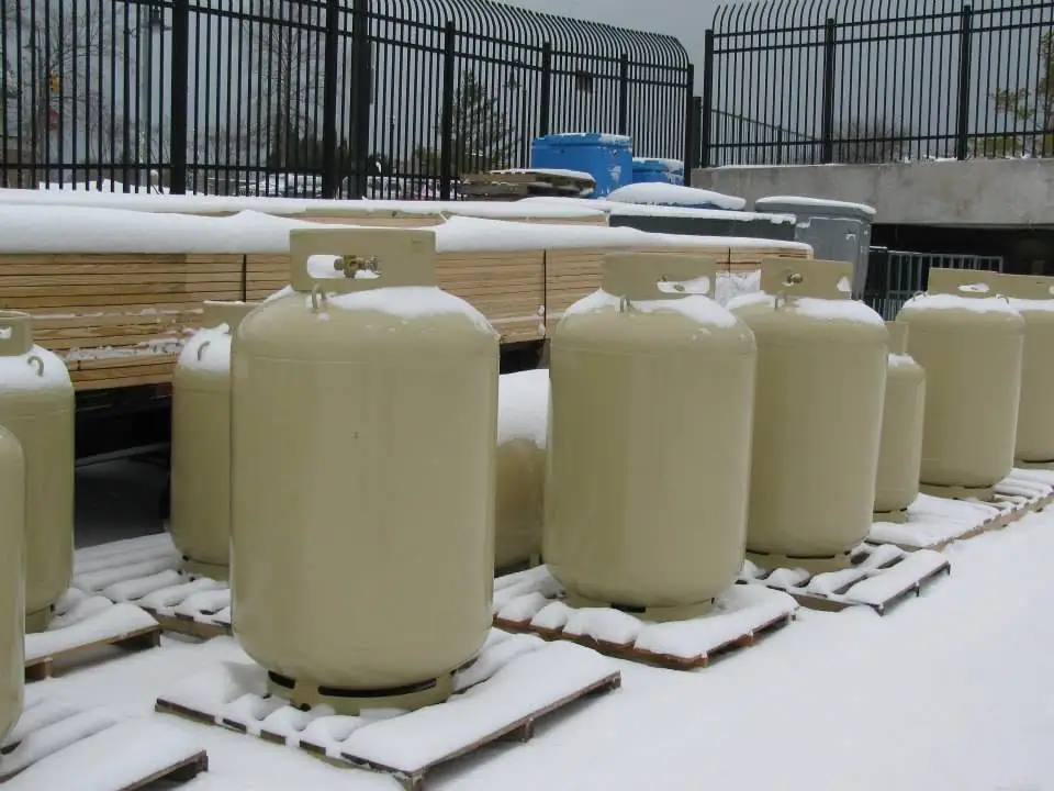 Large cream-colored gas cylinders stand on snow-covered wooden pallets behind a black metal fence.