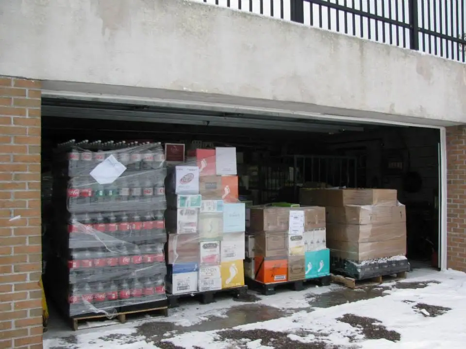 An open garage bay holds pallets of bottled drinks and cardboard boxes beside snow-covered pavement.