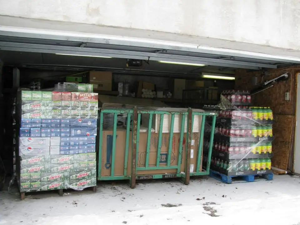 Open garage filled with stacked soft-drink cases and a green hand truck, with snow covering the ground outside.