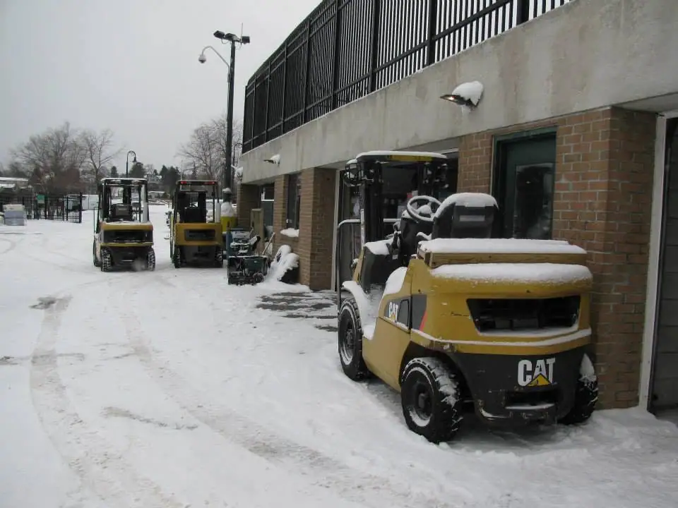 Three snow-covered yellow forklifts are parked beside a brick building in an outdoor service area.