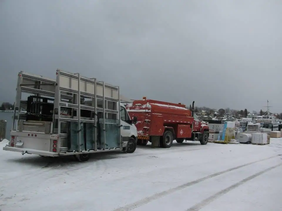 A tall-sided trailer and red tanker fire truck sit on a snowy dock beside a ferry.