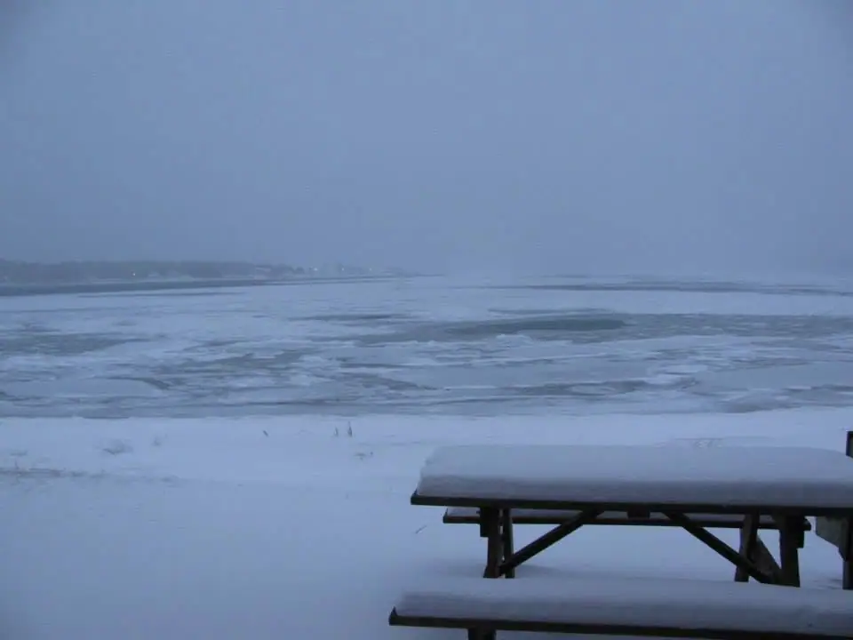 Snow-covered picnic tables overlook broken ice and dark water beneath a gray winter sky.