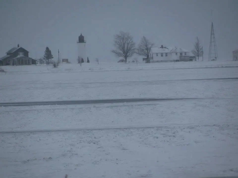 Snow-covered grounds with a lighthouse, houses, bare trees, and a tall lattice tower beneath a gray sky.